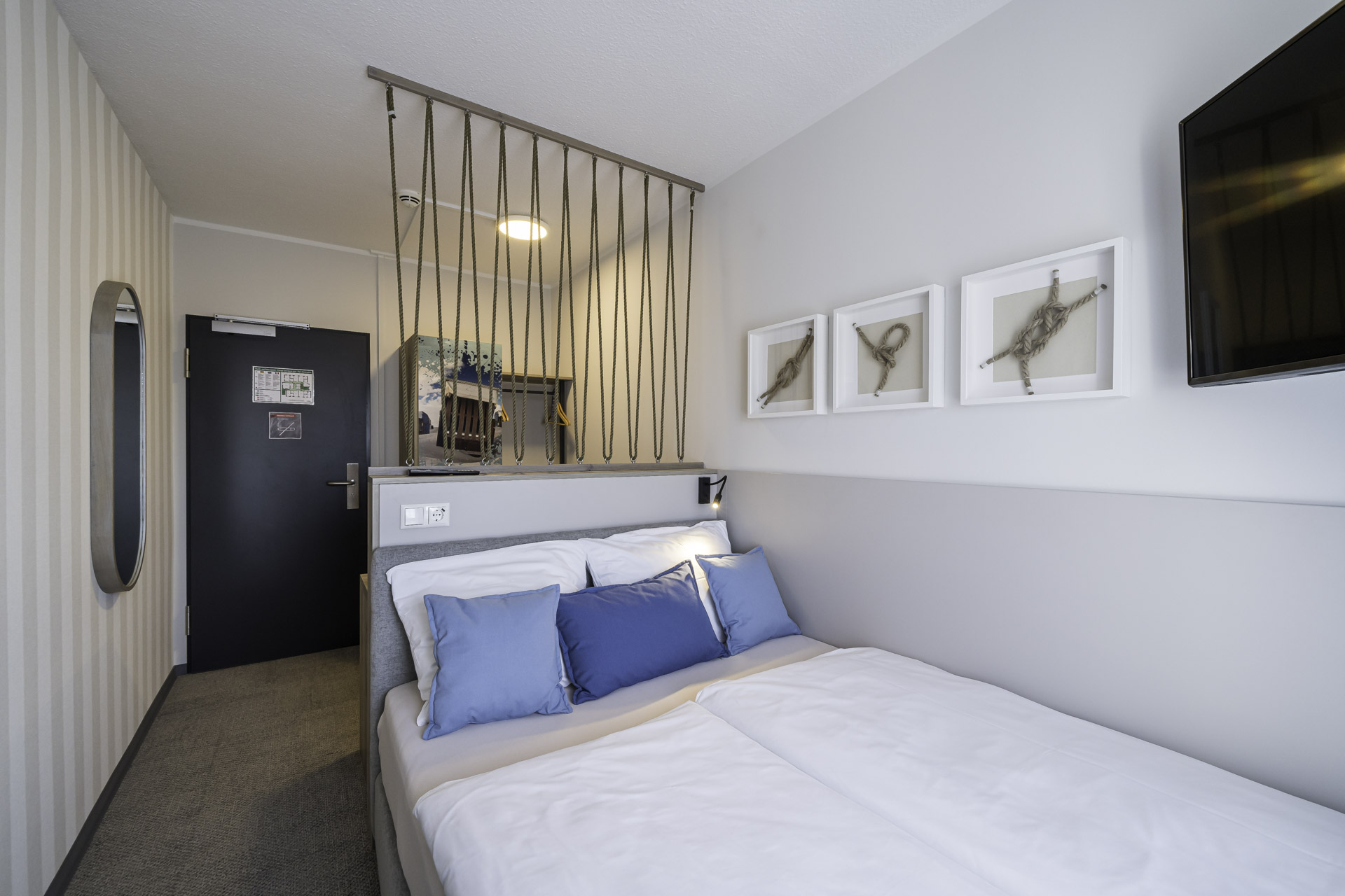 Kleines Doppelzimmer des WIROtels mit Bett, Nachttischen und einer offenen Abtrennung zum Eingangsbereich