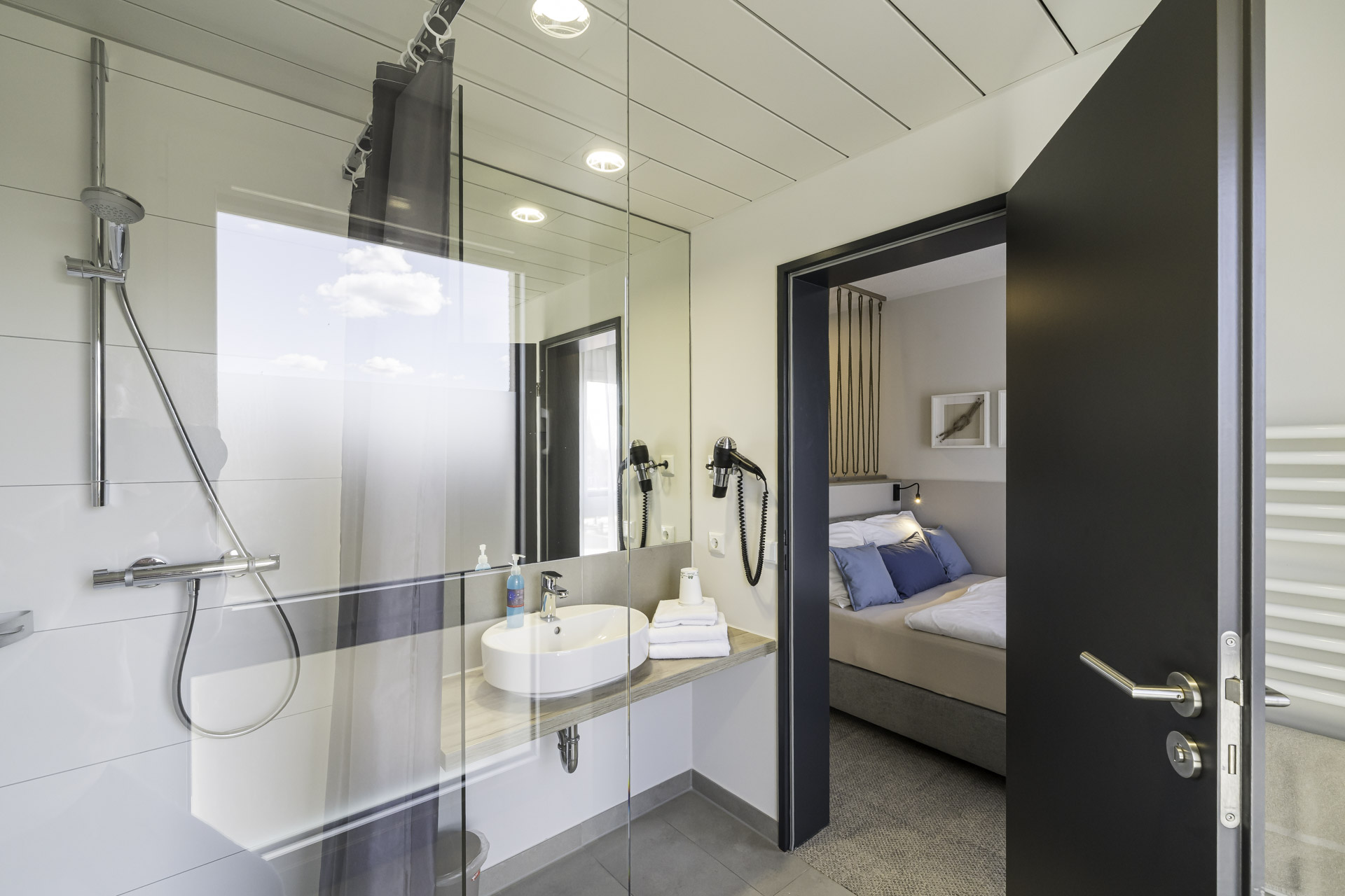 Helles Badezimmer eines kleinen Doppelzimmers mit Dusche, Waschbecken und Blick in den Schlafbereich