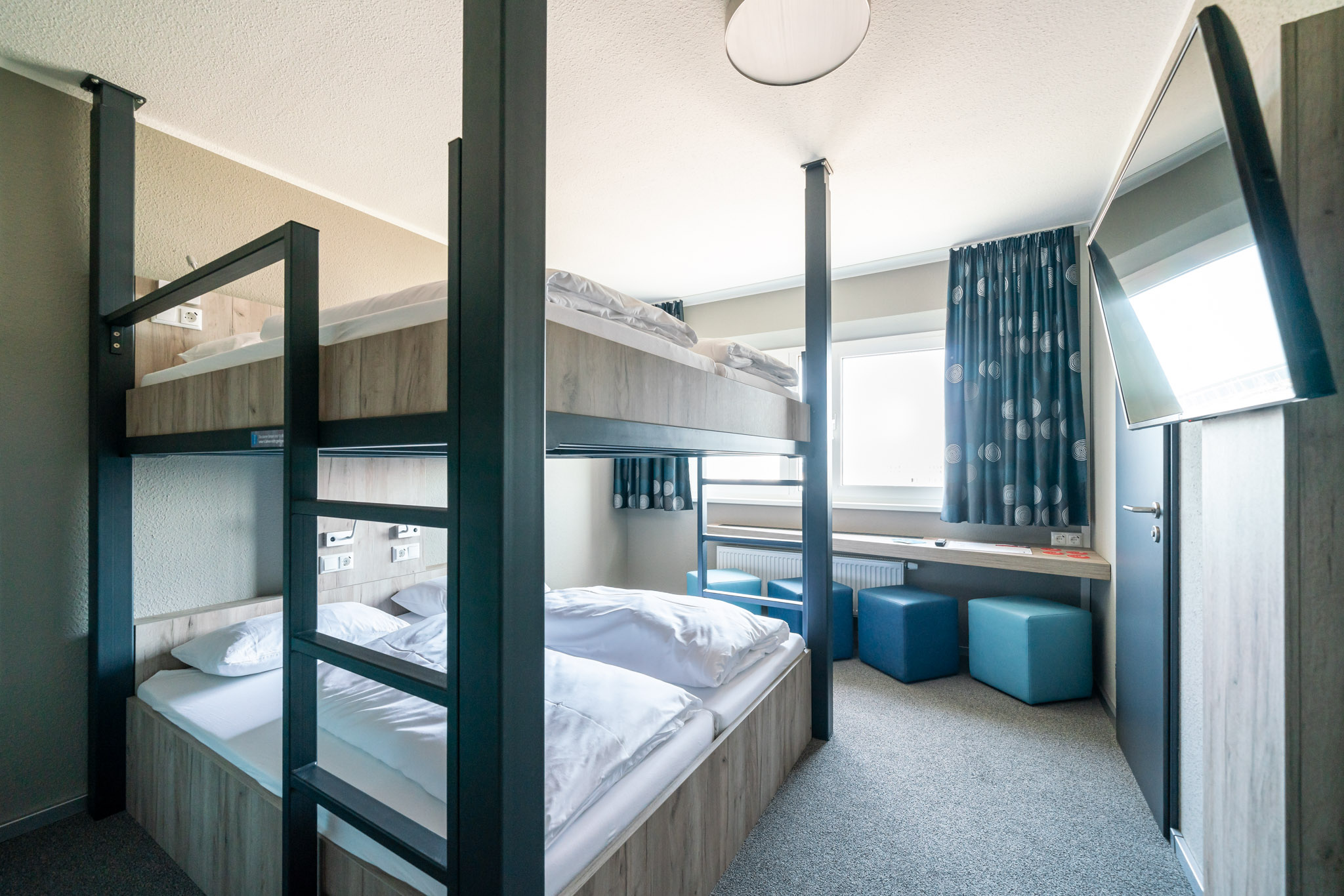 Vierbettzimmer mit zwei Etagenbetten und blauen Hockern am Fenster